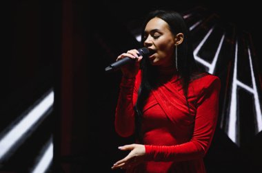 Kadın şarkıcı mikrofonla sahne ışıkları altında şarkı söylüyor, kırmızı bir elbise giyiyor, bir konser etkinliğinde seyircileri eğlendiriyor.