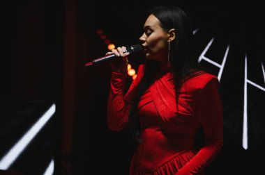 Genç kadın vokalist mikrofon tutuyor, konser sırasında canlı şarkı söylüyor, canlı kırmızı bir kıyafet giyiyor.