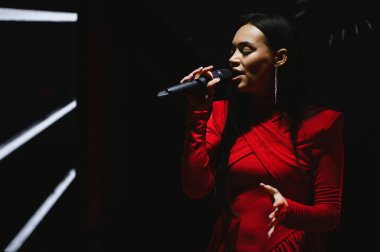 Elinde mikrofon tutan genç bir kadın vokalist, sahne ışıklarıyla karanlık arka planda konser veriyor. Duyguları ve enerjiyi yakalamak