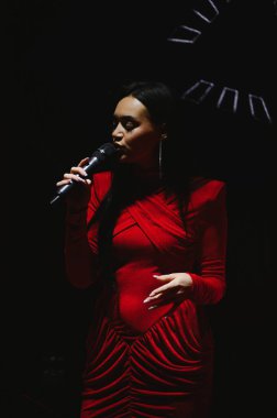 Kadın şarkıcı sahnede şarkı söylüyor, kırmızı bir elbise giyiyor ve spot ışıklarının altında mikrofon tutuyor.