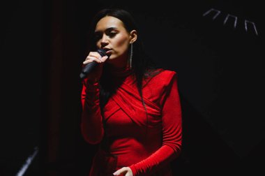 Kadın vokalist mikrofon tutuyor, sahne ışıkları altında sahne alıyor, zarif kırmızı elbiseler giyiyor.