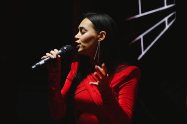 Canlı konser sırasında mikrofonla duygusal şarkı söyleyen kadın şarkıcı spot ışıkları altında kırmızı elbise giyiyor.