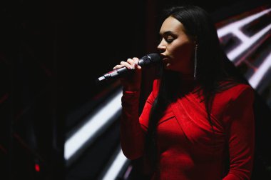 Loş bir sahnede mikrofona şarkı söyleyen genç bir kadın. Kırmızı elbisesiyle onu aydınlatan spot ışıkları.