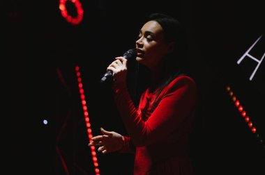 Kırmızı elbiseli genç bir kadın, karanlık bir sahnede şarkı söylüyor, vokal yeteneğiyle seyircileri eğlendiriyor.