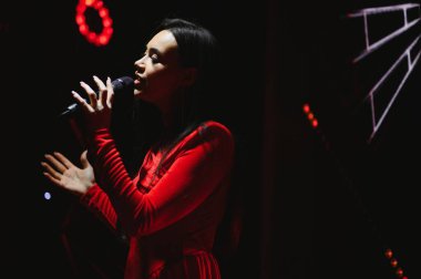 Sahnede kırmızı elbiseli ve mikrofona şarkı söyleyen genç bir vokalist. Sahne ışıkları, duyguları yansıtıyor.