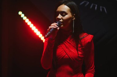 Kadın vokalist konser sahnesinde canlı performans sergiliyor, kırmızı ışıklar altında mikrofona şarkı söylüyor, canlı bir atmosfer yaratıyor.