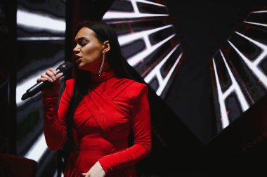 Kırmızı elbiseli, mikrofonlu bir kadın şarkıcı, karanlık bir sahnede grafik ışık desenleriyle canlı müzik yapıyor.