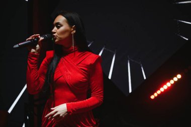 Kırmızı elbiseli kadın vokalist canlı bir konser sırasında dramatik bir sahne ışığı altında mikrofona tutkuyla şarkı söylüyor.