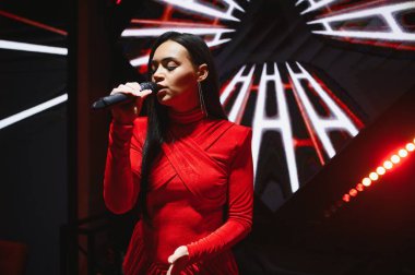 Sahnede elinde mikrofonla dikilen bir kadın, arka planı desenli ve kırmızı ışıklarla şarkı söylüyor.