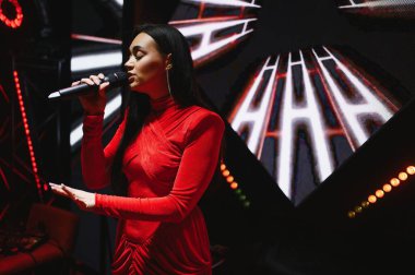 Kırmızı elbiseli genç bir kadın sahnede mikrofona şarkı söylüyor. Canlı müzik performansı yaratıyor.