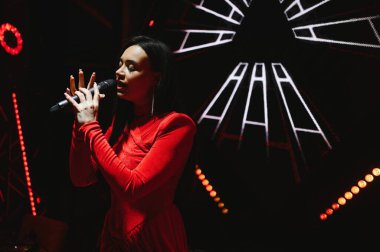 Sahnedeki bir mikrofona tutkuyla şarkı söyleyen kadın sanatçı, kırmızı ışıklar ve soyut arka plan görselleriyle aydınlatılıyor.