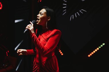 Genç Asyalı kadın mikrofona şarkı söylüyor, sahnede parlak bir sahne ışığı altında güçlü bir müzik performansı sergiliyor.