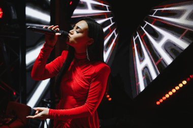 Kırmızı elbiseli kadın vokalist karanlık bir sahnede mikrofonla canlı performans sergiliyor.