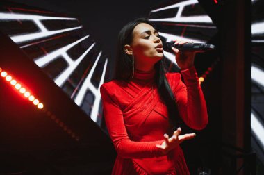 Kırmızı kadife elbiseli genç bir kadın bir müzik konseri veya etkinliği sırasında mikrofona tutkuyla şarkı söylüyor.