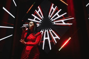 Kırmızı giyinmiş ve elinde mikrofonla sahne ışıkları ve soyut şablonlarla çevrili kadın şarkıcı sesi dinleyicileri büyülüyor.