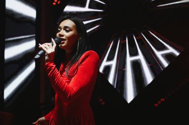 Kırmızı elbiseli genç kadın tek başına şarkı söylüyor. Karanlık bir sahnede mikrofona şarkı söylüyor.