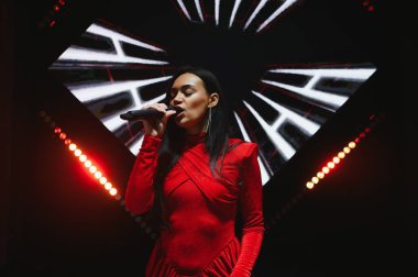 Genç şarkıcı canlı performans sergiliyor, mikrofonu açık sahnede tutuyor. Kırmızı bir elbise giyip güçlü kırmızı ve beyaz ışıklar altında