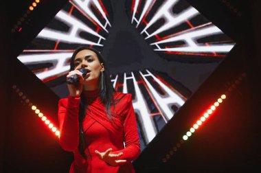 Kırmızı elbiseli kendine güvenen bir kadın, müzik etkinliği sırasında, elinde soyut aydınlatılmış arka planda bir mikrofon tutuyor.