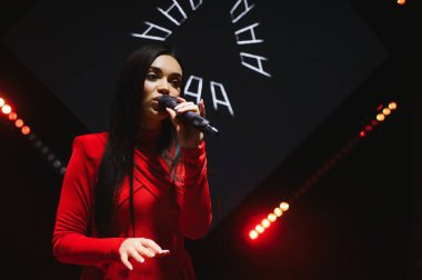Kadın şarkıcı konser sahnesinde canlı performans sergiliyor, şarkı söylerken mikrofon tutuyor. Müzik ve sanatsal performansa odaklanma