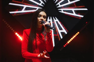 Kırmızı elbise giyen ve sahne ışıklarıyla aydınlatılan bir konser sırasında sahnede şarkı söyleyen kadın şarkıcı.