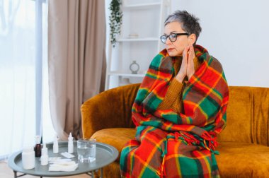 Yetişkin bir kadın grip semptomlarından rahatsız, battaniyeye sarılmış, oturma odasında bir kanepede oturuyor.