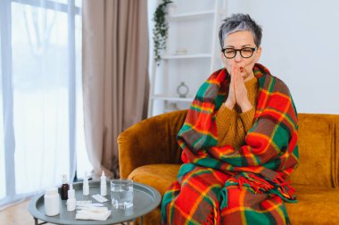 Kıdemli kadın titriyor, sıcak bir battaniyeye sarılmış, ellerini ısıtmaya çalışıyor, evde grip ve hastalık geçiriyor.