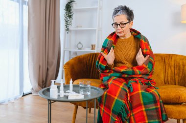 Sıcak bir battaniyeye sarılmış, kanepede oturan yaşlı kadın hastalık ve hastalıktan dolayı titriyor ve kendini iyi hissetmiyor.