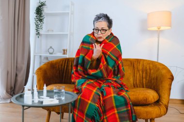 Yaşlı kadın soğuk algınlığı ya da gripten muzdarip, kanepeye sarılı, masanın yanında ilaçla