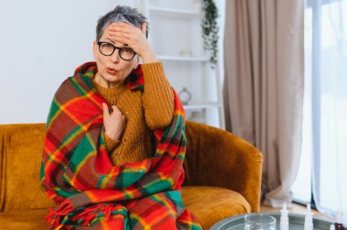 Ateşli ve baş ağrısı olan yaşlı kadın evdeki kanepede battaniyeye sarılmış, dinleniyor ve iyileşme sürecinde huzur arıyor.