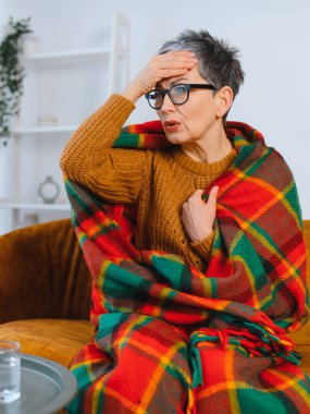 Sıcak bir battaniyeye sarılmış bir kadın baş ağrısı çekiyor ve hasta hissediyor. Yan masada bir bardak su.