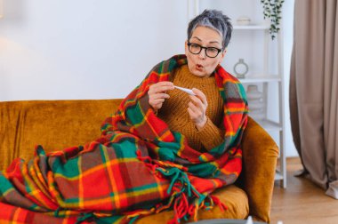 Olgun bir kadın battaniyeye sarılı olarak koltukta oturuyor, termometreye bakarken şaşırmış bir yüz ifadesi takınıyor, üşüyor, ateşi var.