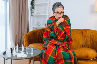 Yaşlı kadın, evde ilaçla çevrili bir şekilde battaniyeye sarılı ve öksürük ve grip belirtileriyle kendini kötü hissediyor.