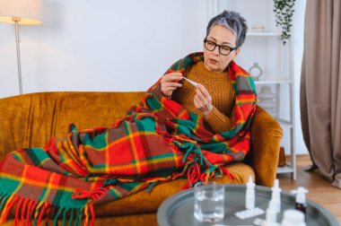 Kareli battaniyeye sarılı, kanepeye oturmuş termometreyle ateşini ölçen, soğuktan hasta hisseden olgun bir kadın.