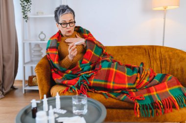Yaşlı kadın üşütmüş ve üşütmüş. Evdeki kanepede battaniyeye sarılıp termometreyi kontrol ediyor.