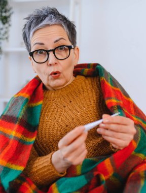 Soğuk algınlığı ve grip belirtileri gösteren yaşlı bir kadın battaniyeye sarılıp vücut ısısını dijital termometreyle ölçüyor.