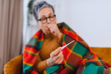 Yaşlı bir kadın battaniyeye sarılmış, termometreyi tutuyor ve öksürürken ağzını kapatıyor, hastalık belirtileri gösteriyor.