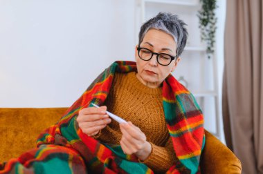 Sıcak ekose battaniyeye sarılı hasta son sınıf öğrencisi bir kadın termometreyle ateşini ölçüyor, grip belirtileri gösteriyor ve evde ateşi var.