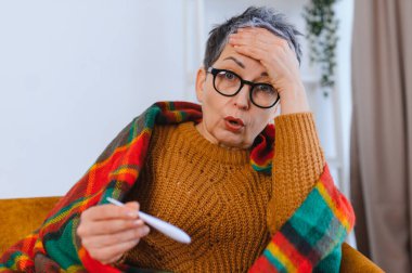 Kadın kendini iyi hissetmiyor, baş ağrısı varken termometreyi kontrol ediyor, hastalık nedeniyle battaniyeye sarılıyor.