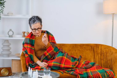 Yaşlı kadın, kanepede battaniyeye sarılıp burun spreyi kullanan grip belirtileriyle kendini kötü hissediyor.