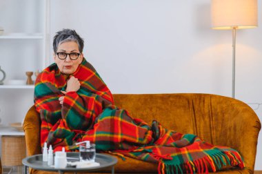 Yaşlı kadın soğuk algınlığı ya da gripten muzdarip. Kanepenin üzerinde ekose bir battaniyeyle, masada ilaçla.