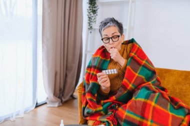 Hastalıktan muzdarip yaşlı bir kadın, battaniyeye sarılmış, nezle ve grip belirtilerini azaltmak için hap alıyor.