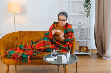 Ekoseli battaniyeyle kaplanmış, kanepeye oturmuş, hastalıktan kurtulmak için bir paket hap tutan olgun bir kadın.