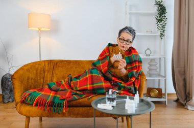 Nezle ya da soğuk algınlığı belirtileri gösteren yaşlı bir kadın. Kanepeye sarılmış, su toplamış bir paketten ilaç alıyor.