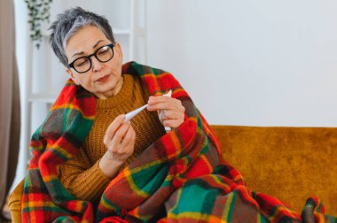 Yaşlı kadın üşütmüş ya da grip olmuş. Kanepede battaniyeye sarılı halde vücut ısısını ölçüyor.