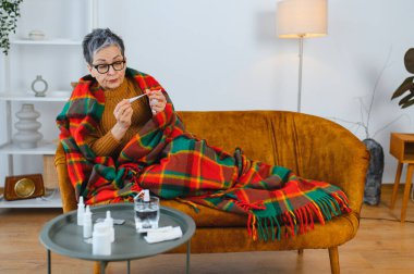 Yaşlı kadın soğuk algınlığı geçiriyor, kanepede battaniyeye sarılıp ateşini ölçüyor.