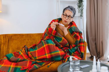 Üst düzey kadın, battaniyeye sarılmış, termometreyle ateşini ölçüyor, soğuk algınlığıyla ya da evde grip belirtileriyle mücadele ediyor.