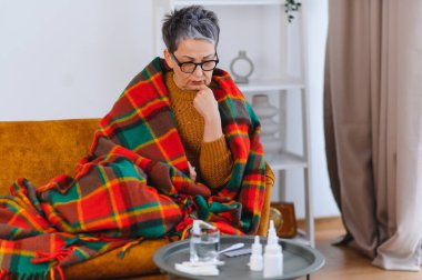 Yaşlı kadın kendini iyi hissetmiyor, battaniyeye sarılmış, kanepeye oturmuş, yanında bir bardak su ve ilaçla.