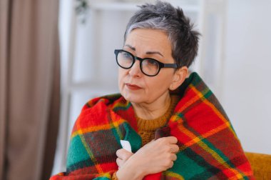 Yaşlı kadın kendini hasta hissediyor, battaniyeye sarılmış, nezle ya da grip belirtileri gösteriyor, elinde mendil tutuyor.