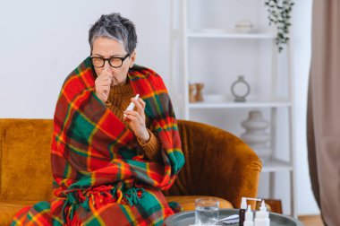 Yaşlı kadın soğuk algınlığı ve grip yüzünden kendini kötü hissediyor. Burnunu siliyor ve sıcak bir battaniyeye sarılı halde burun spreyi kullanıyor.