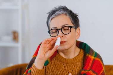 Soğuk algınlığı, grip ya da mevsimsel alerji belirtilerini azaltmak için burun spreyi kullanan bir kadın battaniyenin altında ısınıyor.